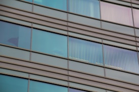 Glass windows of a modern building with horizontal metal frames and reflective surfaces. Some windows show vertical blinds partially drawn, allowing a view of interior silhouettes.