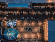 An aerial view of a parking lot featuring a large blue and white striped tent resembling a circus tent. Numerous cars are parked in a grid-like arrangement, and the scene is illuminated by streetlights, creating a dynamic contrast with shadows. There are buildings on the top and sides of the image.