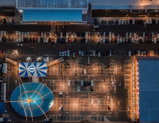 An aerial view of a parking lot featuring a large blue and white striped tent resembling a circus tent. Numerous cars are parked in a grid-like arrangement, and the scene is illuminated by streetlights, creating a dynamic contrast with shadows. There are buildings on the top and sides of the image.