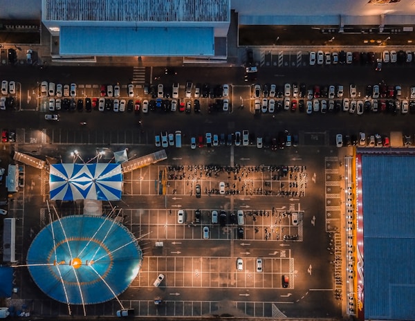 An aerial view of a parking lot featuring a large blue and white striped tent resembling a circus tent. Numerous cars are parked in a grid-like arrangement, and the scene is illuminated by streetlights, creating a dynamic contrast with shadows. There are buildings on the top and sides of the image.