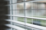 Horizontal blinds partially open with soft natural light filtering through. The focus is on the slats, while the background remains blurred giving a sense of depth.