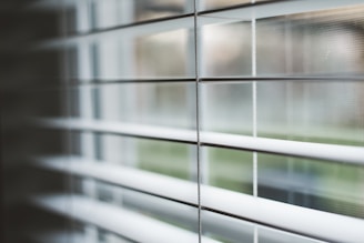 Horizontal blinds partially open with soft natural light filtering through. The focus is on the slats, while the background remains blurred giving a sense of depth.