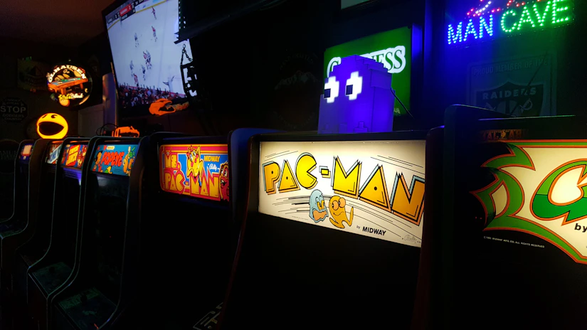 A row of classic arcade machines with colorful displays, including well-known games like Pac-Man and Ms. Pac-Man. Neon lights adorn the scene with a 'Man Cave' sign visible. A large screen in the background shows a sports game, adding to the lively ambiance.