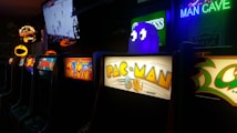A row of classic arcade machines with colorful displays, including well-known games like Pac-Man and Ms. Pac-Man. Neon lights adorn the scene with a 'Man Cave' sign visible. A large screen in the background shows a sports game, adding to the lively ambiance.