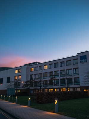 A modern office building with large windows illuminated from inside, set against a twilight sky that transitions from blue to pink. The foreground features a well-maintained pathway lined with small lights, and a lawn with neat hedges. The building has a sign with the letters 'wbs' on the facade.
