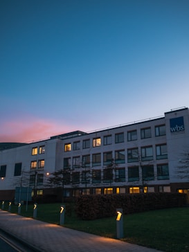 A modern office building with large windows illuminated from inside, set against a twilight sky that transitions from blue to pink. The foreground features a well-maintained pathway lined with small lights, and a lawn with neat hedges. The building has a sign with the letters 'wbs' on the facade.