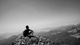 A solitary figure sitting quietly on a mountain ridge at sunrise, gazing thoughtfully into the distance.