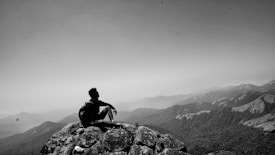 A person is sitting alone on a rocky mountain peak, gazing over a vast landscape of rolling hills and mountains beneath a hazy sky. The scene conveys solitude and contemplation.