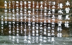 White Japanese text is painted on a dark wooden background. The text is arranged in vertical lines and occupies a significant portion of the surface. The wood appears aged and weathered, enhancing its rustic appearance.