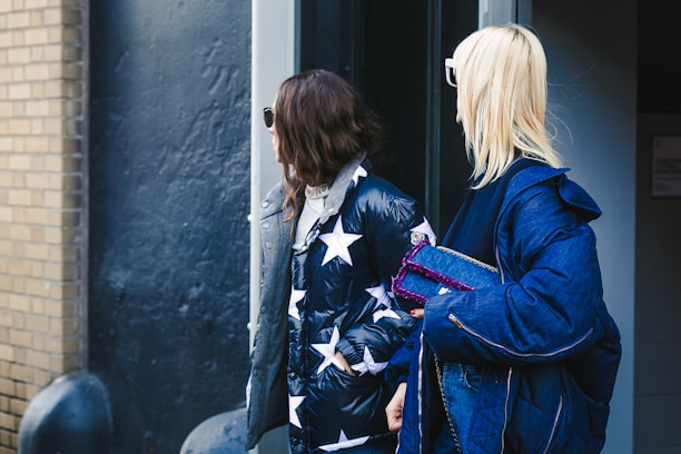 Two individuals dressed in stylish winter attire are standing outdoors. One person is wearing a puffy jacket with star patterns, while the other is in a denim jacket. They both have sunglasses and appear to be in a casual urban setting.