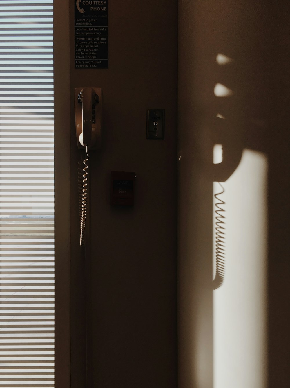 white corded telephone mounted on wall near window with window blinds