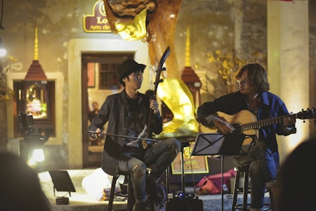 Two musicians are performing indoors, each playing a different instrument. One musician is playing a traditional string instrument while wearing a dark jacket and hat. The other musician is playing an acoustic guitar, wearing a blue shirt. The setting appears to be warm and intimate, with subdued lighting and decorative elements in the background, such as a golden sculpture and framed artwork.