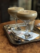 Two cocktail glasses filled with a creamy beige drink are placed on a metal tray on a wooden table. Small purple flower petals decorate the top of the drinks. Accompanying the drinks, there is a teaspoon and a sugar shaker on the tray.