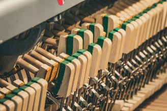 A detailed close-up of piano hammers and strings, showcasing the intricate mechanism. The hammers have a wooden frame with green and yellow felt pads at the striking surface, and metal wires and connections are visible.