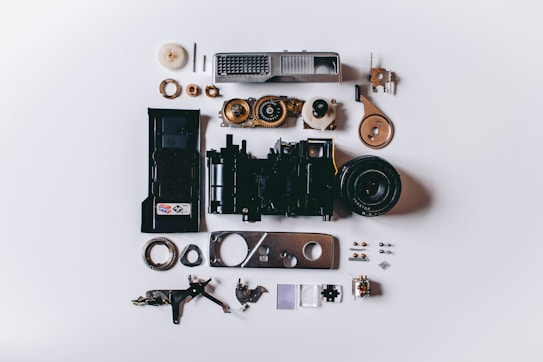 An array of various disassembled camera parts is neatly arranged on a flat surface. The components include gears, a lens, screws, casings, and electronic parts, showcasing the intricate details and inner workings of a camera.