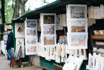An outdoor art stall displays a variety of paintings and prints, with many depicting urban and scenic landscapes. The stall is situated in a shaded area with trees in the background. A person wearing a dark hat and coat stands to the left, seemingly observing the collection. The artworks are hung neatly on racks and are surrounded by other art supplies and materials.