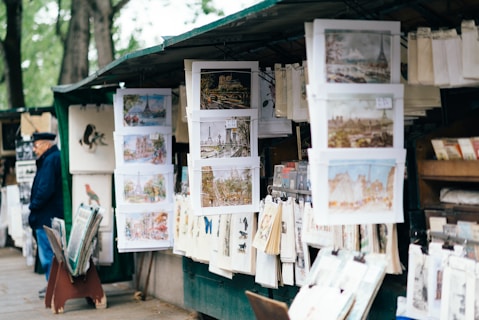 An outdoor art stall displays a variety of paintings and prints, with many depicting urban and scenic landscapes. The stall is situated in a shaded area with trees in the background. A person wearing a dark hat and coat stands to the left, seemingly observing the collection. The artworks are hung neatly on racks and are surrounded by other art supplies and materials.