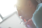 A person is smoking a cigarette, with sunlight illuminating the side of their face. The focus is soft, and smoke is visible near the fingers and mouth, giving a hazy effect.