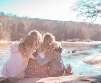 three women sitting on rock near body of water surrounded by trees during daytime