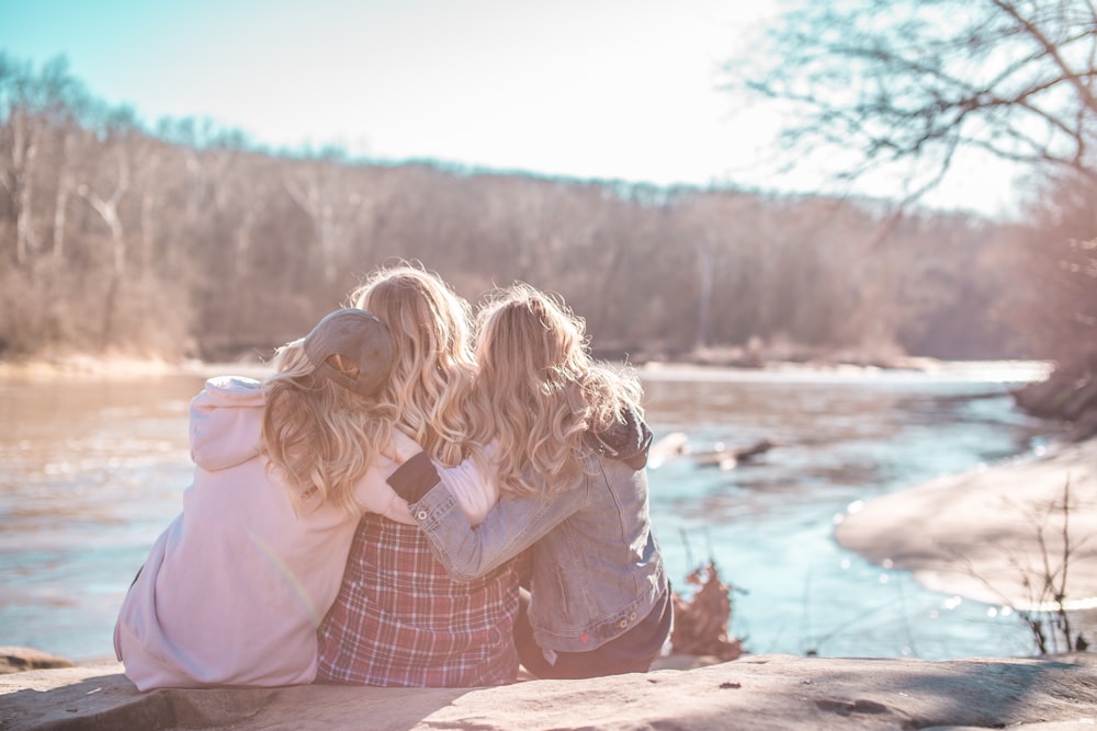 three women sitting on rock near body of water surrounded by trees during daytime