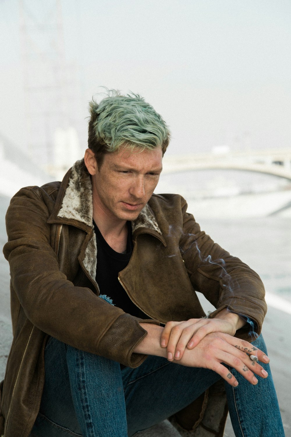 man with green hair in black shirt and brown coat sits on brown sand near white suspension bridge