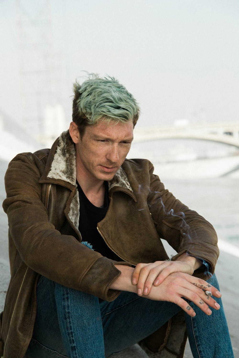 Homem com cabelo verde em camisa preta e casaco marrom senta-se na areia marrom perto da ponte suspensa branca