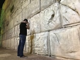 A person stands facing a large stone wall, with detailed carvings on the stones. The individual appears to be engaged in a moment of reflection or prayer, with their head bowed and hands touching the wall. Shadows cast on the wall add depth to the scene.