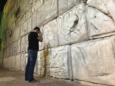 A person stands facing a large stone wall, with detailed carvings on the stones. The individual appears to be engaged in a moment of reflection or prayer, with their head bowed and hands touching the wall. Shadows cast on the wall add depth to the scene.