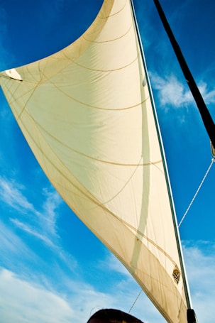 A large white sail is prominently displayed against a clear blue sky. The sail captures sunlight, creating soft shadows and a glowing effect. A few wisps of clouds are visible in the background.