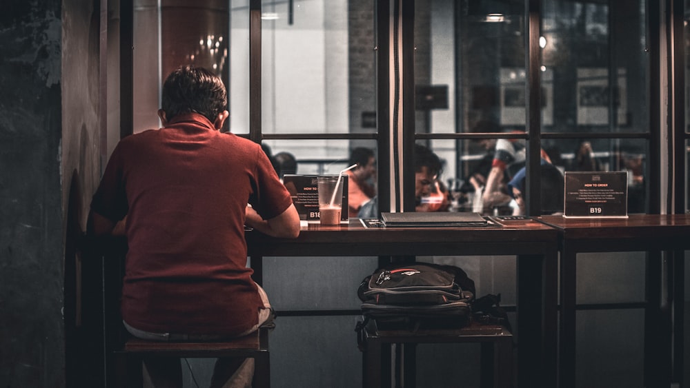 man sitting on brown wooden bar stool beside backpack on wooden bar stool