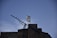 A Greek flag flutters atop a stone building against a clear blue sky. In the foreground, an antenna is visible, pointing diagonally upward.