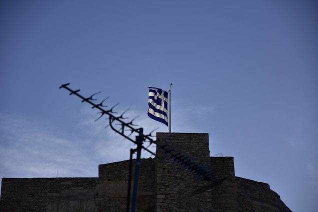 A Greek flag flutters atop a stone building against a clear blue sky. In the foreground, an antenna is visible, pointing diagonally upward.