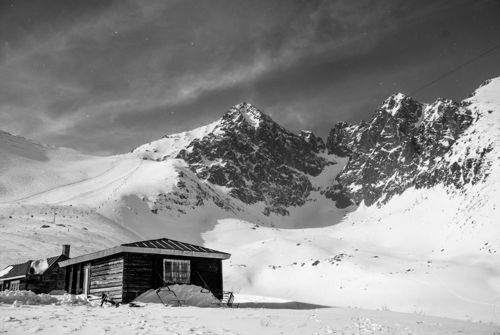 Foto en escala de grises de una casa en la montaña cubierta de nieve