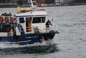 A passenger boat with the name 'Tortur 1' is navigating through choppy waters. Several people are visible on the deck, some seated and others standing. Life rings and safety equipment are mounted on the sides of the vessel. In the background, a cityscape with various buildings can be seen.