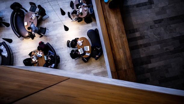 An overhead view of a modern indoor space with people sitting around circular wooden tables. The seating includes dark, curved couches and chairs. Laptops and notebooks are scattered on the tables, indicating a study or working environment. The lighting is subtle, with cone-shaped pendants hanging above.