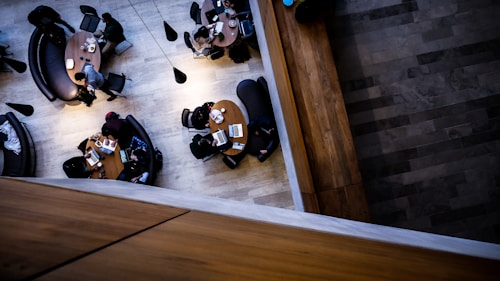 An overhead view of a modern indoor space with people sitting around circular wooden tables. The seating includes dark, curved couches and chairs. Laptops and notebooks are scattered on the tables, indicating a study or working environment. The lighting is subtle, with cone-shaped pendants hanging above.