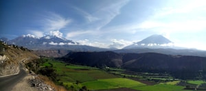 A breathtaking view of a lush green valley bordered by majestic mountains with snow-capped peaks under a clear blue sky. A winding road runs along the side of the mountain, leading into the distance. The landscape is bathed in sunlight, enhancing the vibrant colors and creating distinct shadows.