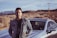 A happy customer standing next to a shiny rental car in a scenic location.