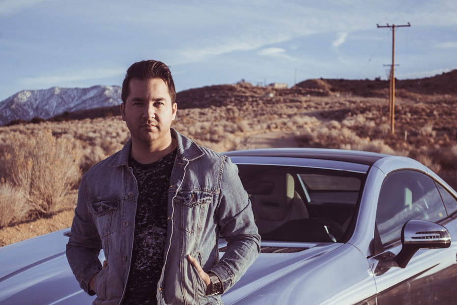 A person stands confidently next to a sleek car in a desert landscape. The car is parked on a dirt road surrounded by dry brush and bushes. There are mountains visible in the background with a clear sky above.