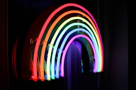 A vibrant neon light in the shape of a rainbow, showcasing a full spectrum of colors against a dark background. The glow from the neon tubes creates a striking contrast, emphasizing each color of the rainbow.