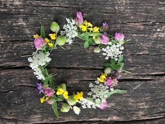 A wreath made of various wildflowers and green foliage is laid out on a weathered wooden surface. The wreath contains a mix of white, yellow, purple, and pink flowers.