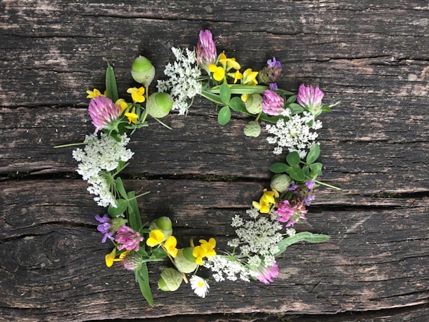 A wreath made of various wildflowers and green foliage is laid out on a weathered wooden surface. The wreath contains a mix of white, yellow, purple, and pink flowers.