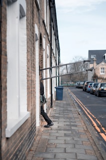 A person is climbing into a residential building through a window with the help of a ladder. The brick building is adjacent to a sidewalk, and several cars are parked along the street. The houses have white window trims, and the street has double yellow lines.
