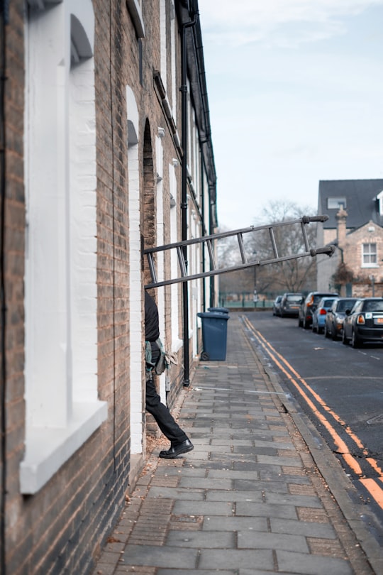 man carrying grey ladder entering brown concrete building during daytime in Cambridge United Kingdom