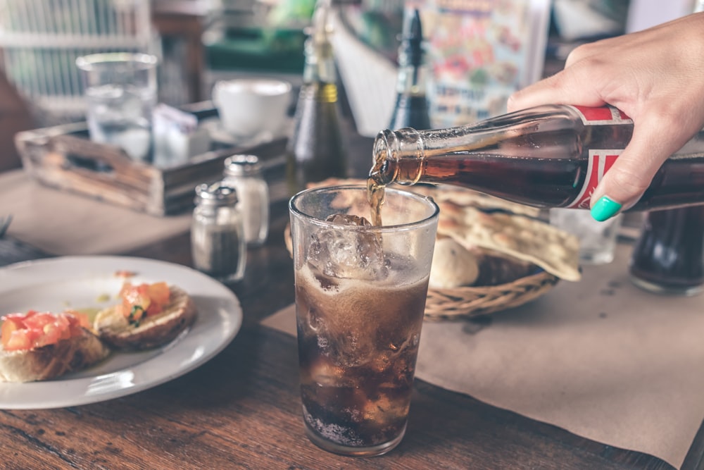 person pouring Coca-Cola soda on clear drinking glass near round white ceramic plate