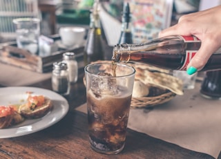 person pouring Coca-Cola soda on clear drinking glass near round white ceramic plate