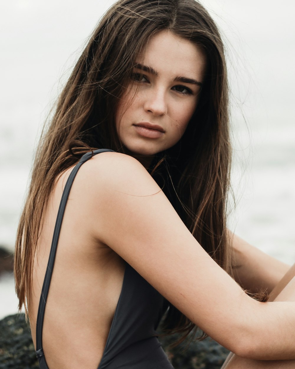 woman black tank swimsuit sits on black rock near body of water posing for photoshoot