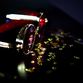 A pair of round sunglasses with reflections on the lenses is placed on a dark surface. Surrounding the sunglasses are small, scattered golden gears or sequins that catch the light, creating a shimmering effect. The lighting is low, emphasizing the contrast between the bright reflective surfaces and the dark background.