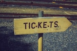 A wooden directional sign with the word 'TICKETS' painted in dark letters. The sign is mounted on a metal pole and set against a background of gravel and railway tracks.