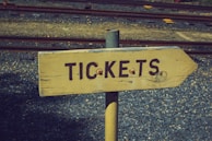 A wooden directional sign with the word 'TICKETS' painted in dark letters. The sign is mounted on a metal pole and set against a background of gravel and railway tracks.